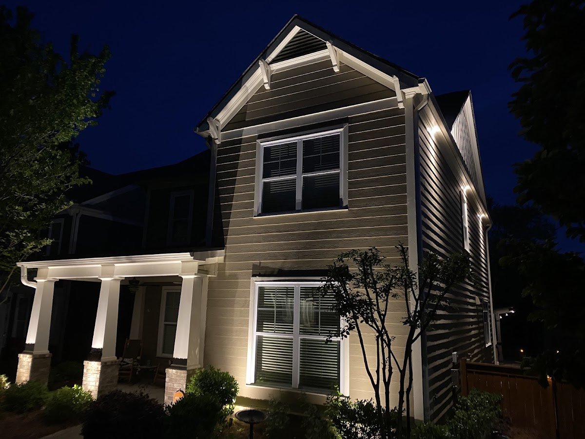 Two-story home at night with warm white landscape uplighting washing the gable, lap siding and front porch columns from below.