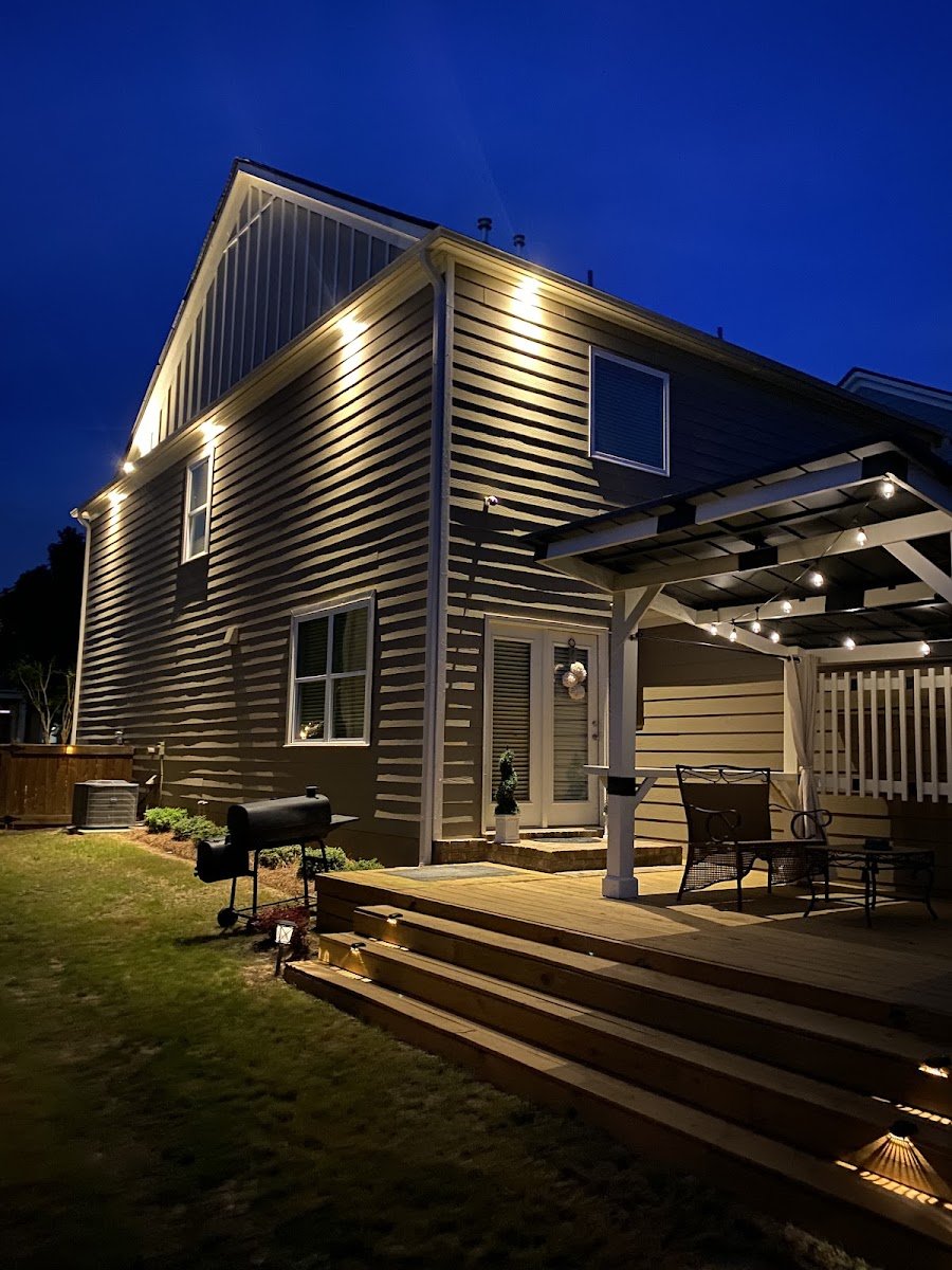Backyard of a two-story home at dusk, soffit downlights on the siding and string lights over a pergola-covered deck with patio furniture.