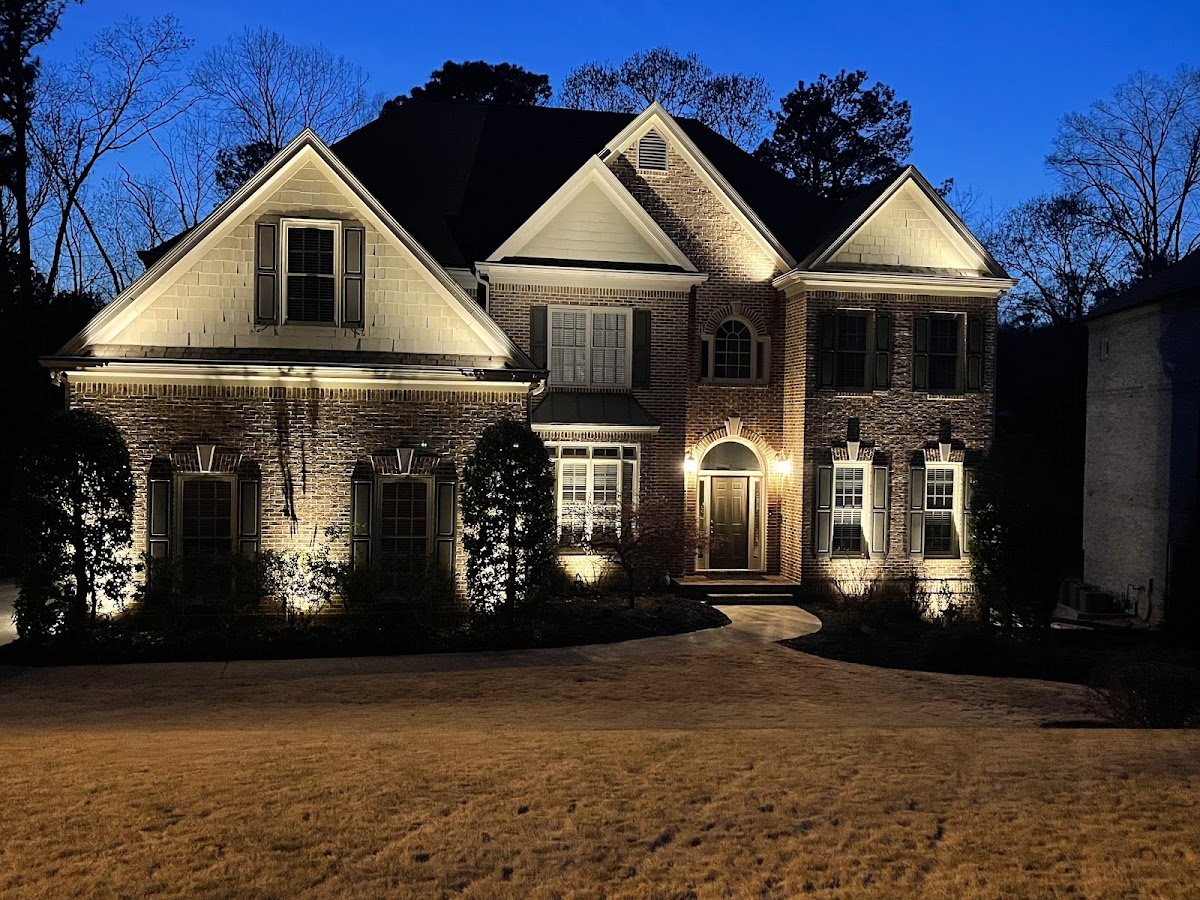 Large two-story brick home at dusk with warm landscape uplighting highlighting the gables, columns and front facade above a curved driveway.