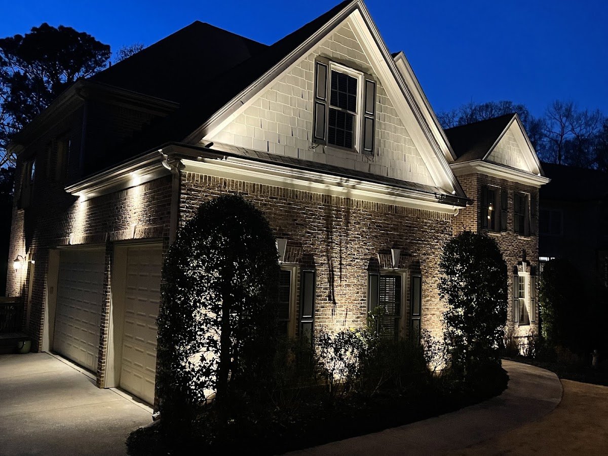 Side of a brick two-story home at dusk with landscape uplighting illuminating the gable, brickwork and manicured shrubs along the driveway.