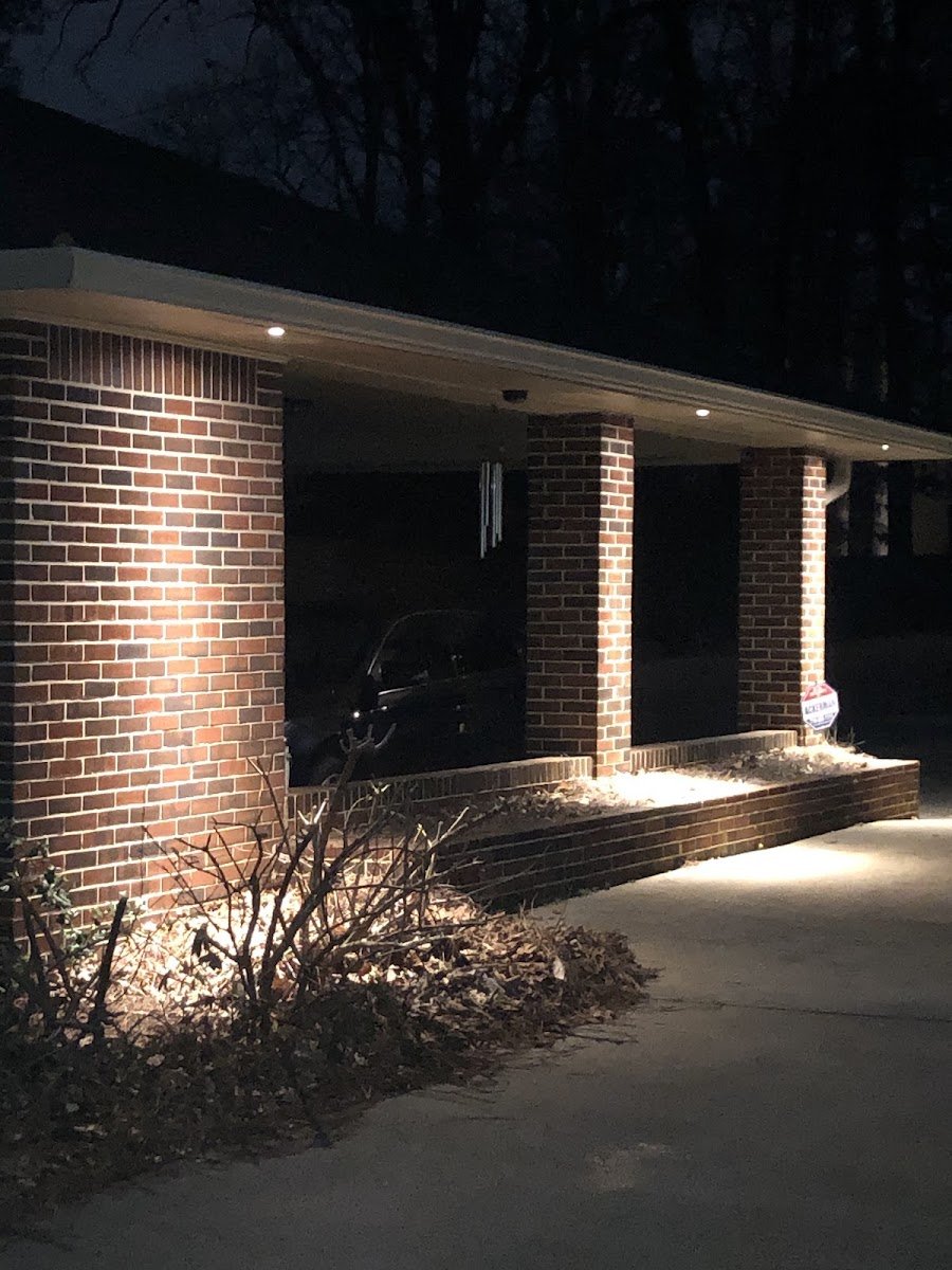 Brick home's covered porch at night with recessed soffit downlights and landscape lighting uplighting the columns and bushes below.