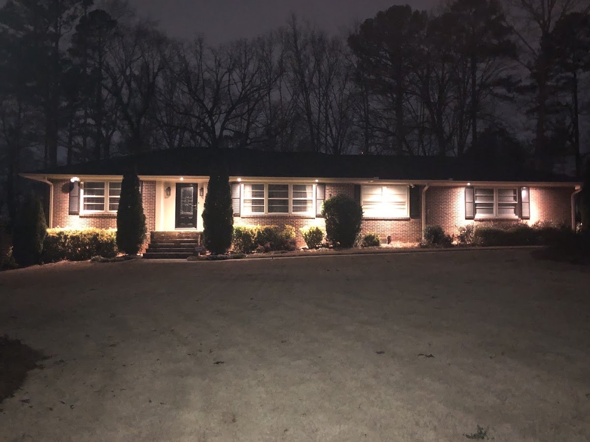 A single-story brick ranch home at night with warm white uplighting washing the facade and shrubs, surrounded by tall bare trees and manicured bushes.