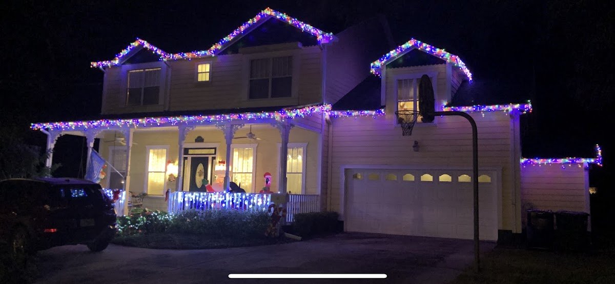 A two-story home outlined at the rooflines and gables with multicolor twinkle lights, wraparound porch and picket fence lit, holiday inflatables in the yard.