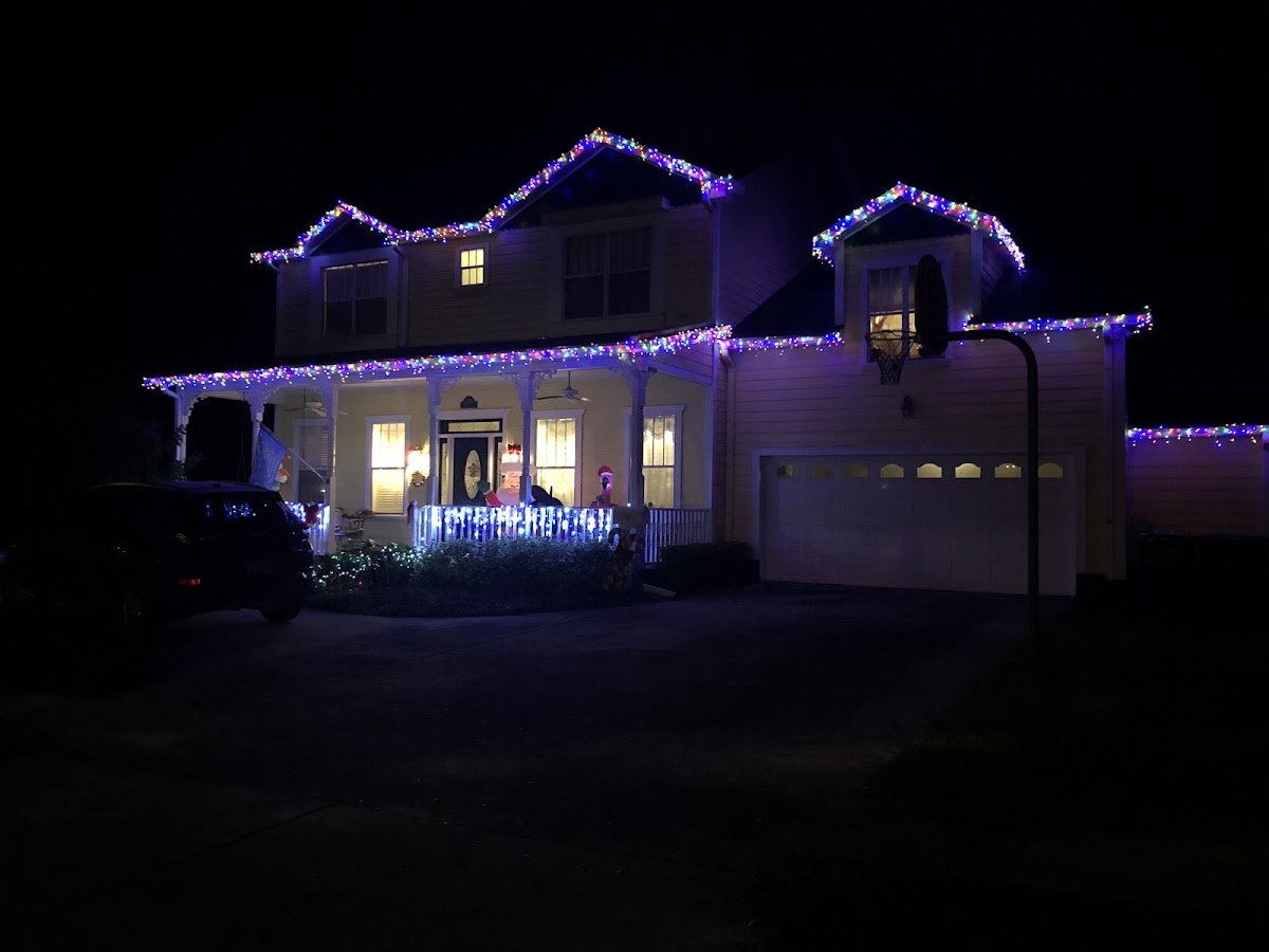 A two-story home with multicolor icicle lights outlining every roofline and gable at night, front porch glowing and blue lights on the porch railing.