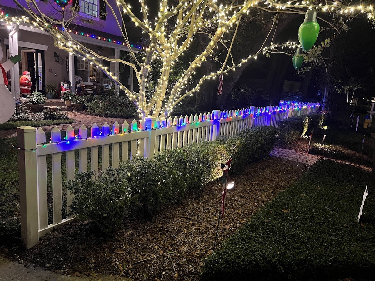 A white picket fence and tree trunk wrapped in warm white mini lights with multicolor bulbs along the fence, holiday figures on the porch beyond.