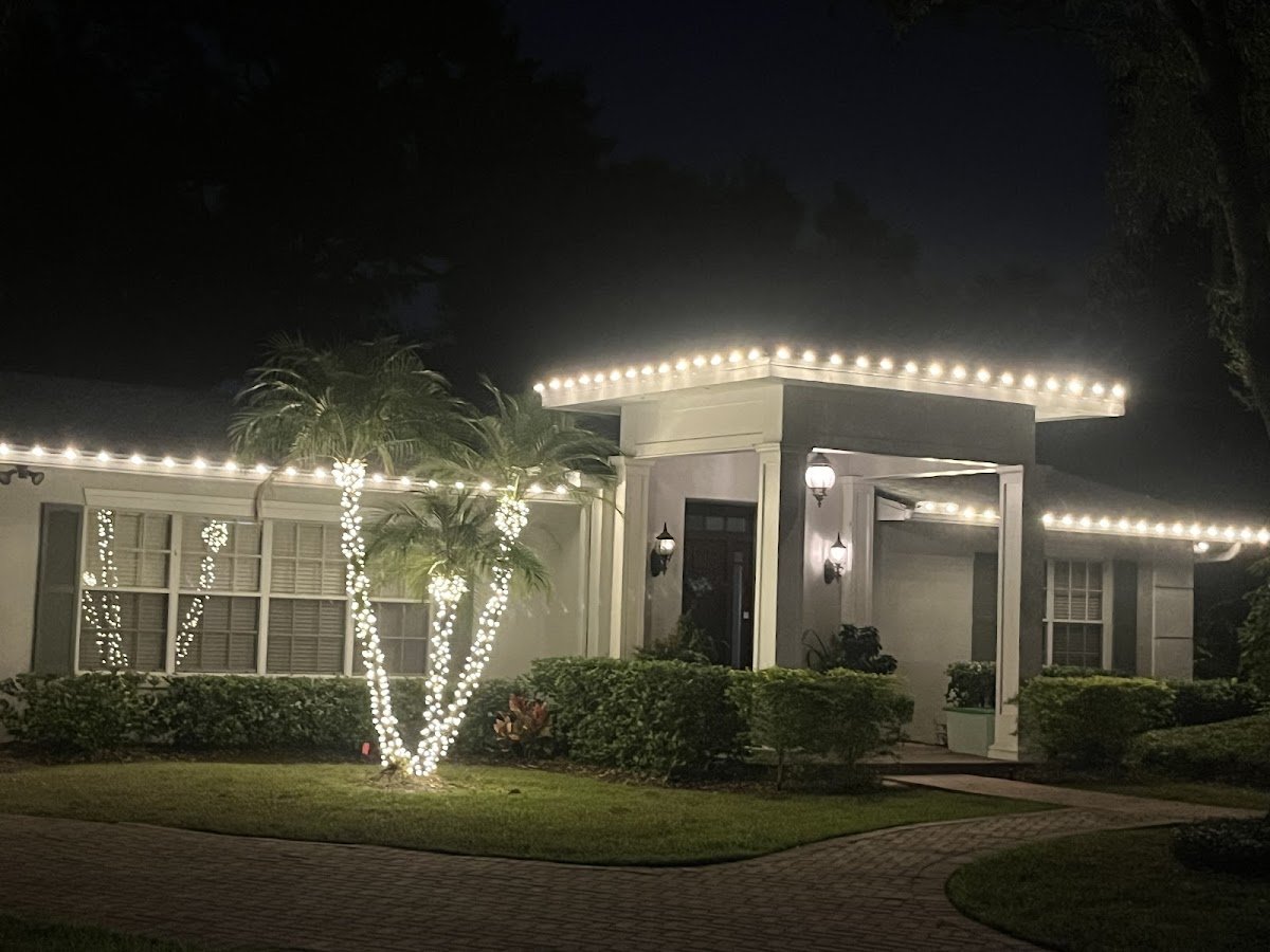 A single-story stucco home outlined in warm white C9 bulbs along the roofline, palm trunks wrapped in mini lights, glowing at night.