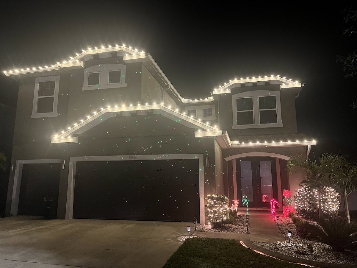 A two-story home with warm white C9 bulbs outlining the peaked rooflines, a lit garage-side wreath and glowing shrubs by the entry at night.