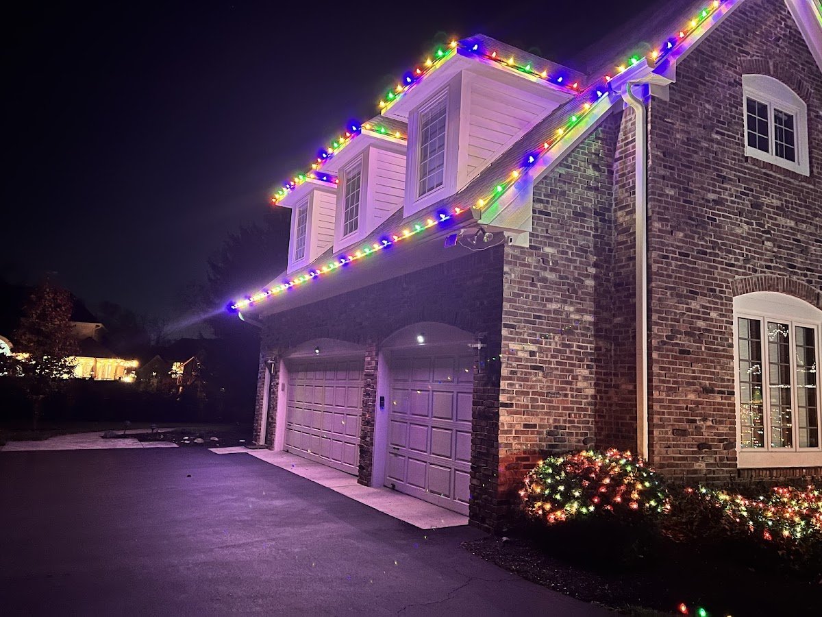 A two-story brick home with multicolor C9 bulbs outlining the rooflines and dormers, garage below and multicolor lights in the front shrubs at night.
