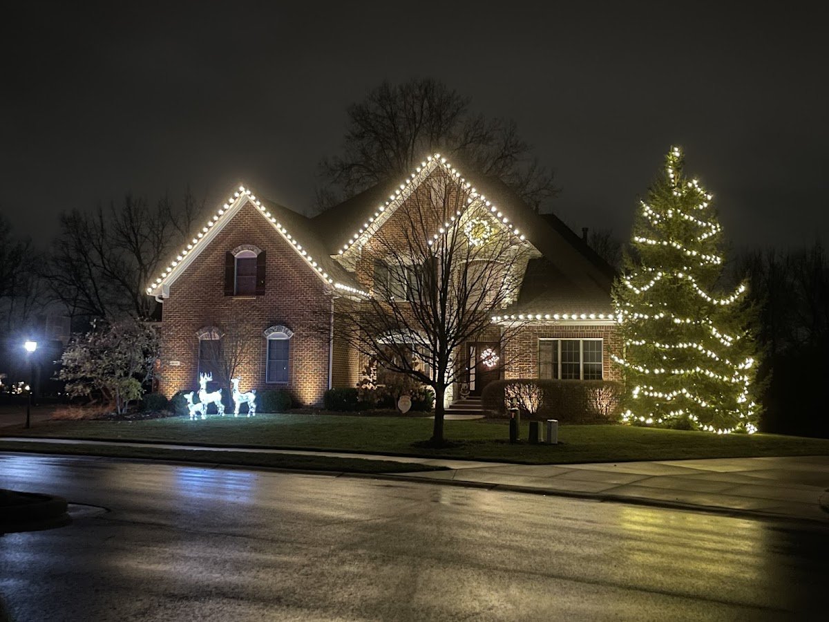 A brick home outlined in warm white C9 roofline lights with a fully lit evergreen tree, lighted reindeer figures and wet street reflecting at night.