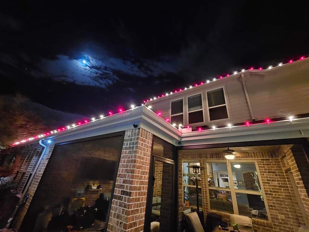 Looking up at the roofline of a brick two-story home outlined in alternating red and white C9 Christmas bulbs, the moon glowing in the night sky.