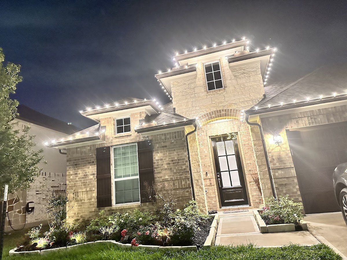A two-story brick and stone home with warm white C9 Christmas lights outlining every roofline and peak, landscaping and front door softly uplit at night.