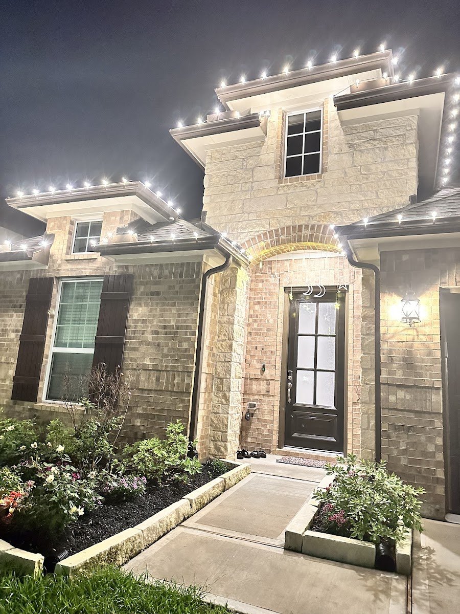Close-up of a brick and stone home's front entry with warm white C9 bulbs tracing the roof edges and gables, glowing over the door at night.