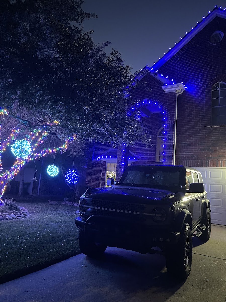 A two-story brick home outlined in blue Christmas lights along the roofline and arch, front-yard trees hung with blue and multicolor lit spheres, Ford Bronco parked at night.
