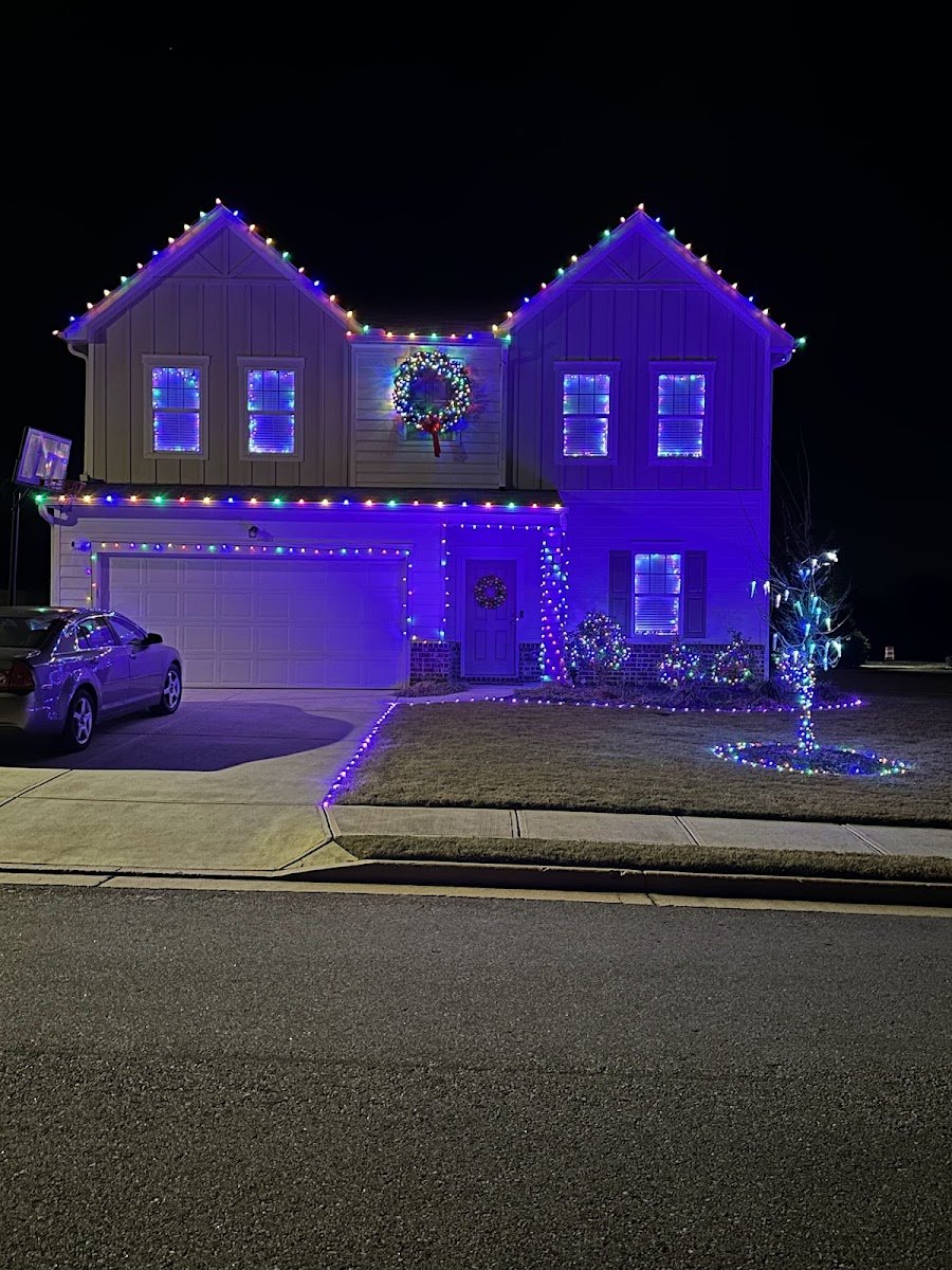 A two-story brick home outlined in warm white C9 lights with tree trunks wrapped in lights, blue-lit shrubs and wreaths, viewed from the street at night.