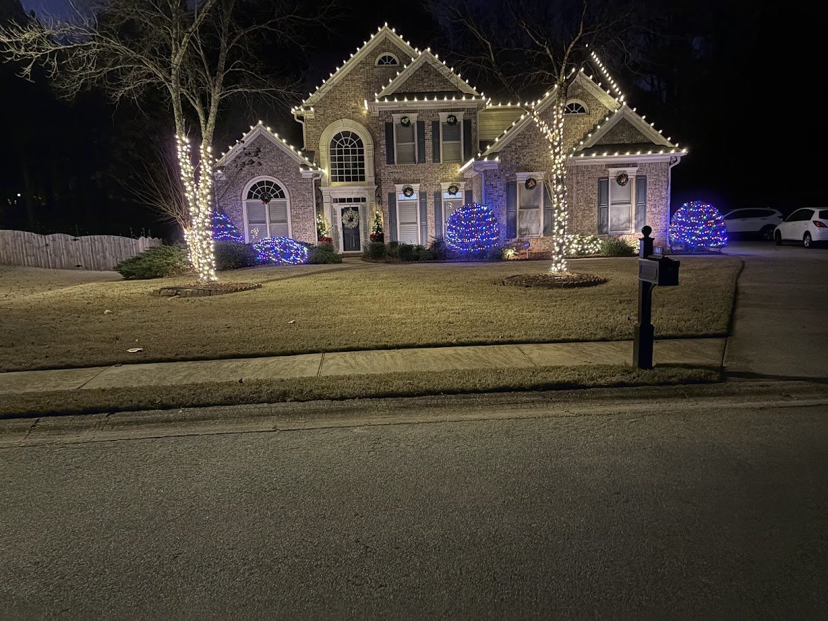A two-story home with lit wreaths in the windows, columns wrapped in string lights and shrubs covered in red, green and white Christmas lights, slightly blurred at night.