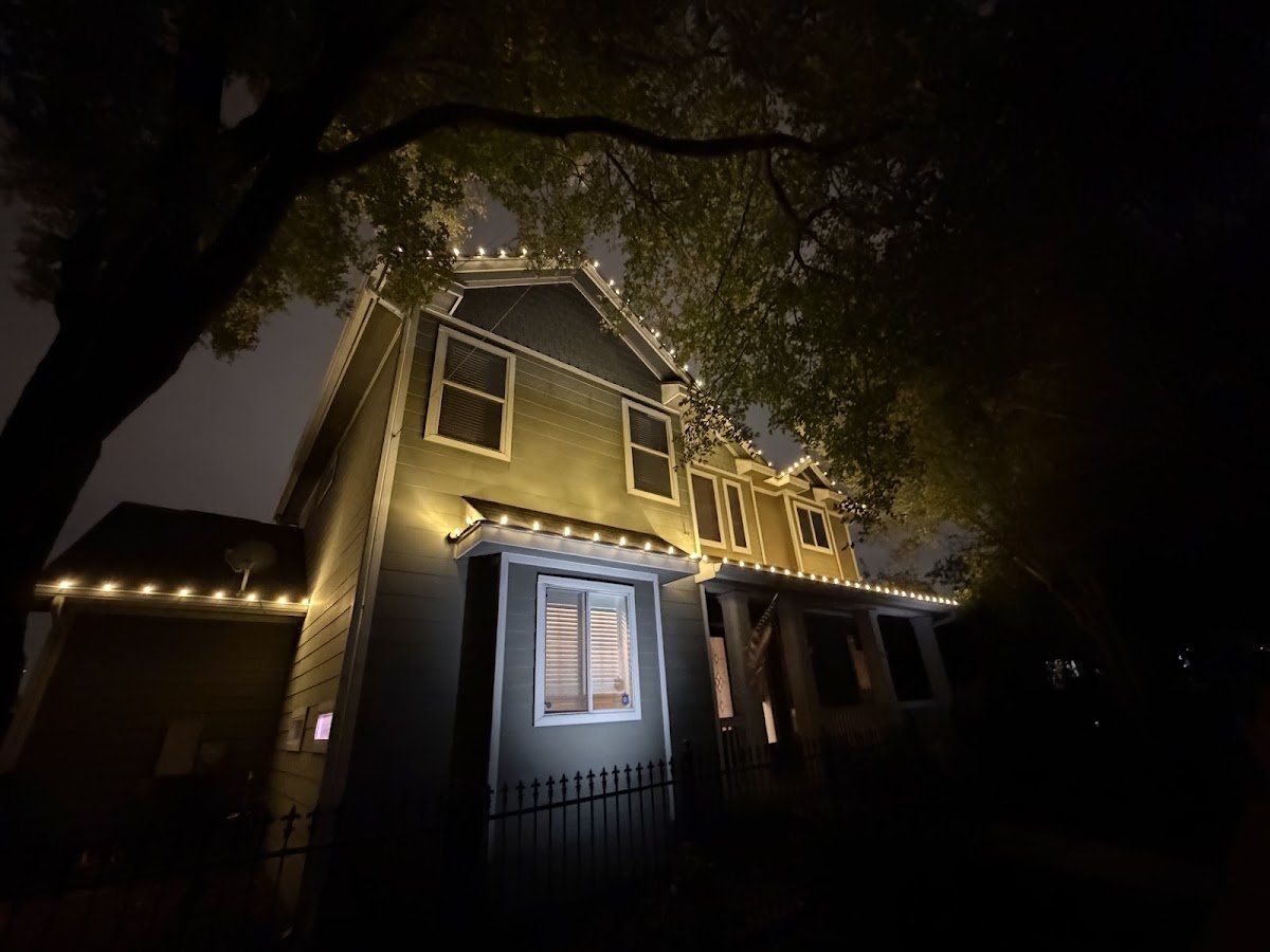 Two-story home at night with warm white lights outlining the roofline and porch, framed by a large overhanging tree and iron fence.