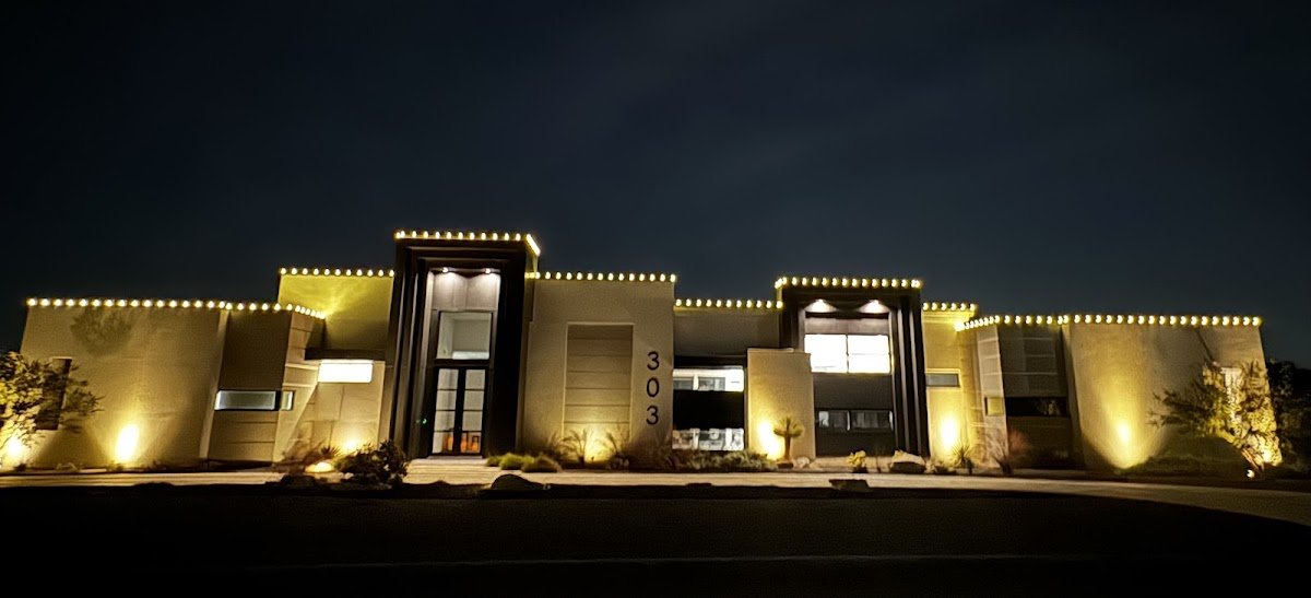 Modern flat-roofed home outlined in warm white roofline lights with amber uplighting washing the stucco walls at night.